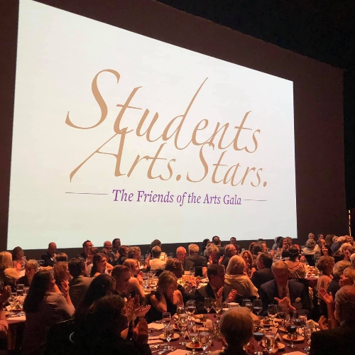 Gala event with many people sitting on a stage looking at a banner for Friends of the Arts that says Students, Arts, Stars