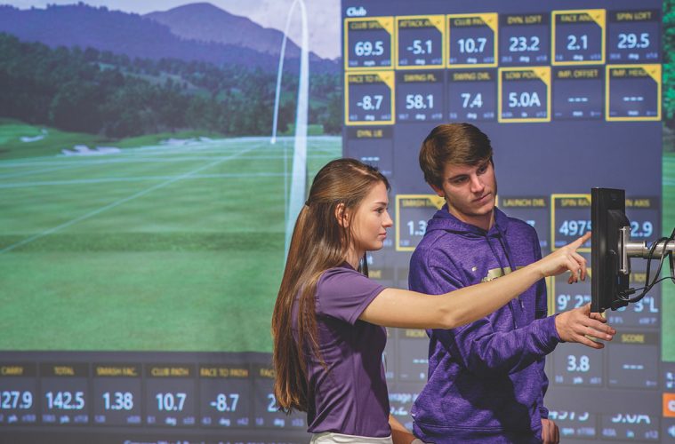 Two people standing shoulder to shoulder, the woman points showing something to her male counterpart. There is a screen with a simulated golf course in the back ground.