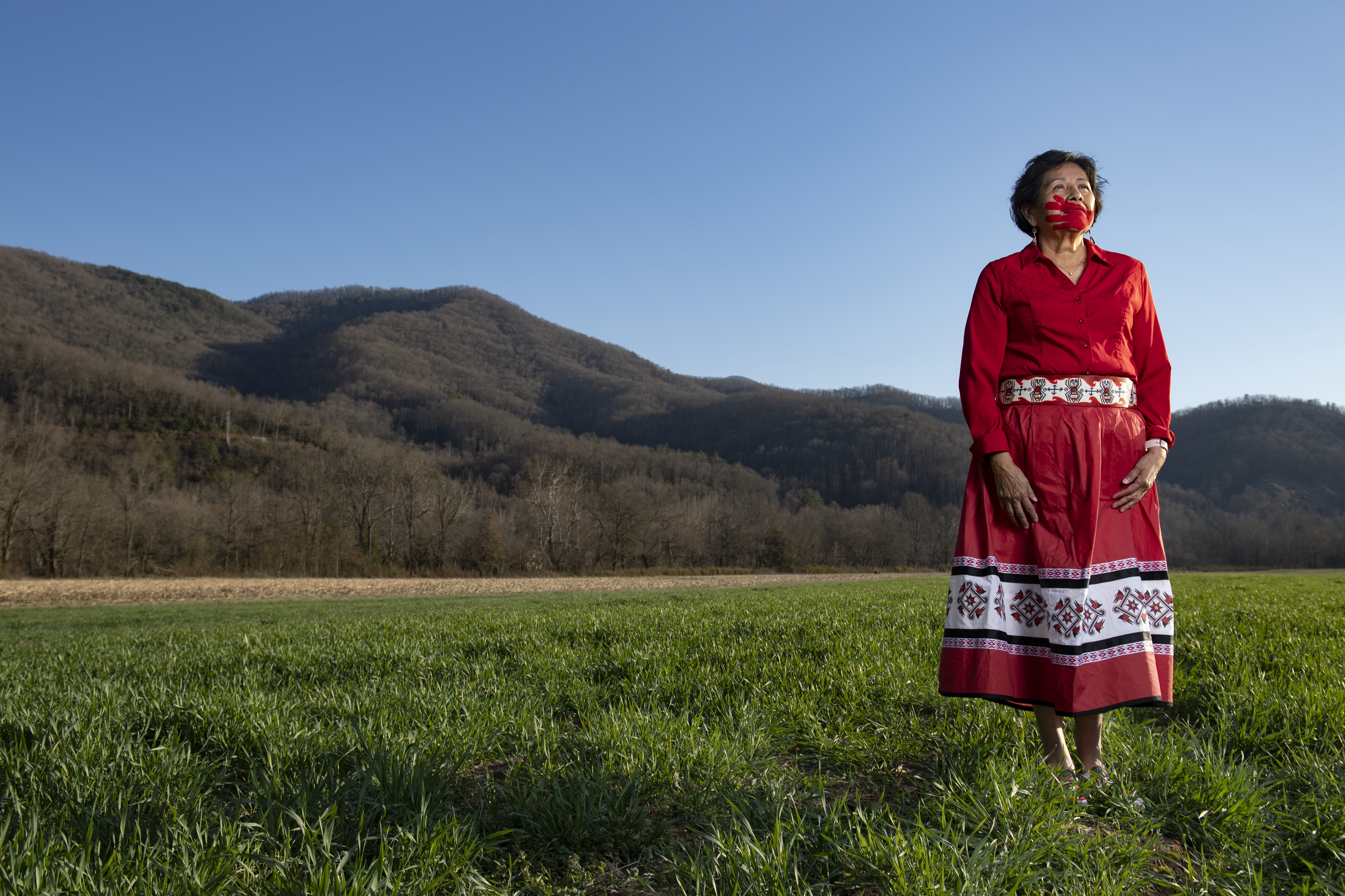 Elder woman standing in a red ribbon dress with a red handprint on her face signifying MMIW