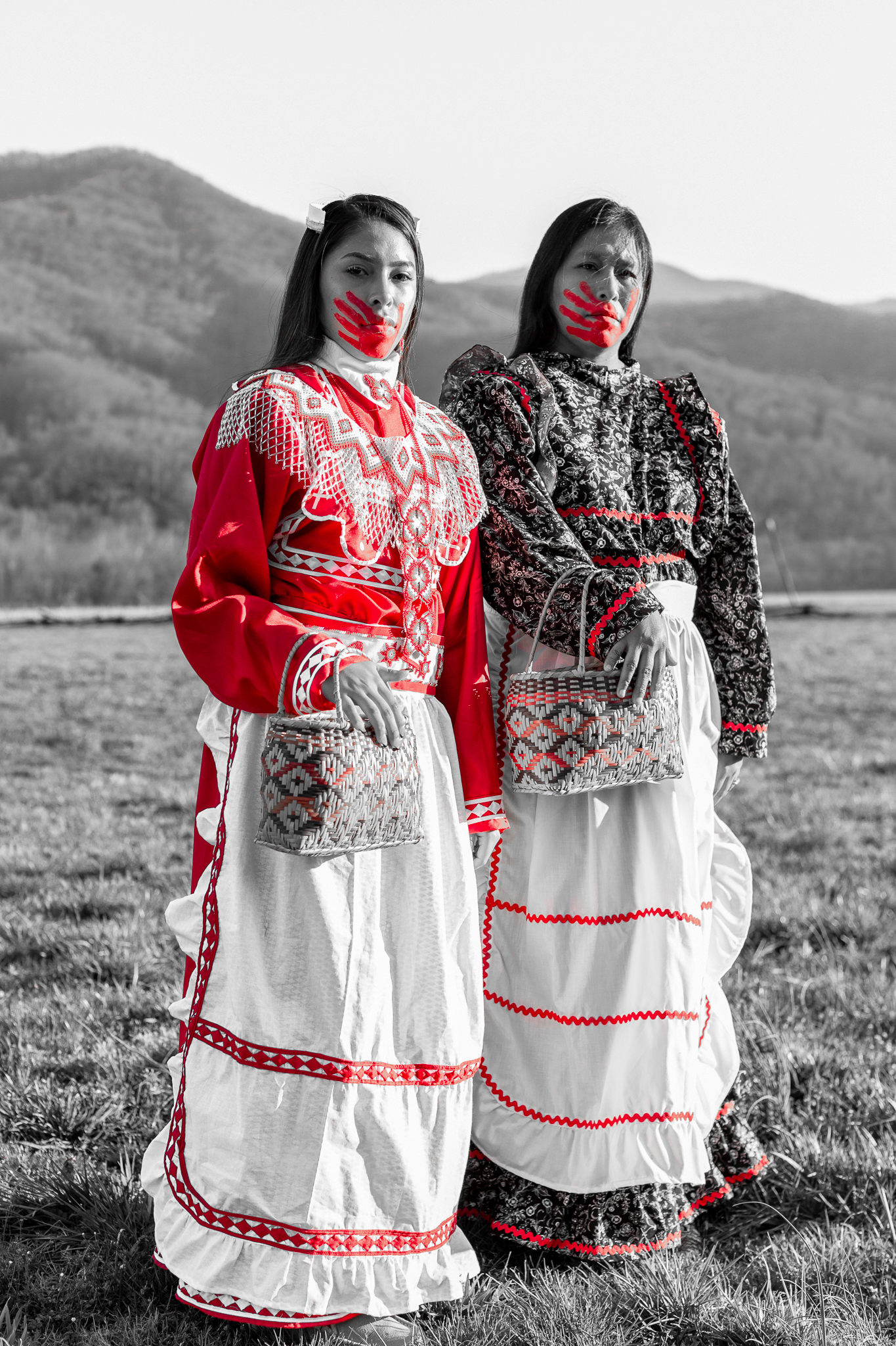 Two indigenous women in ribbon dresses standing in a field with a red handprint over their mouths symbolizing the MMIW movement