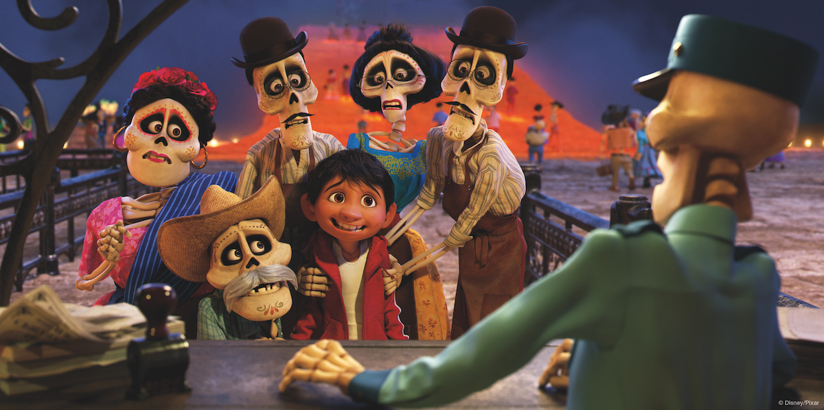 5 skeletons are dressed in traditional mexican clothes. All skeletons are standing around a young boy in a red sweatshirt 