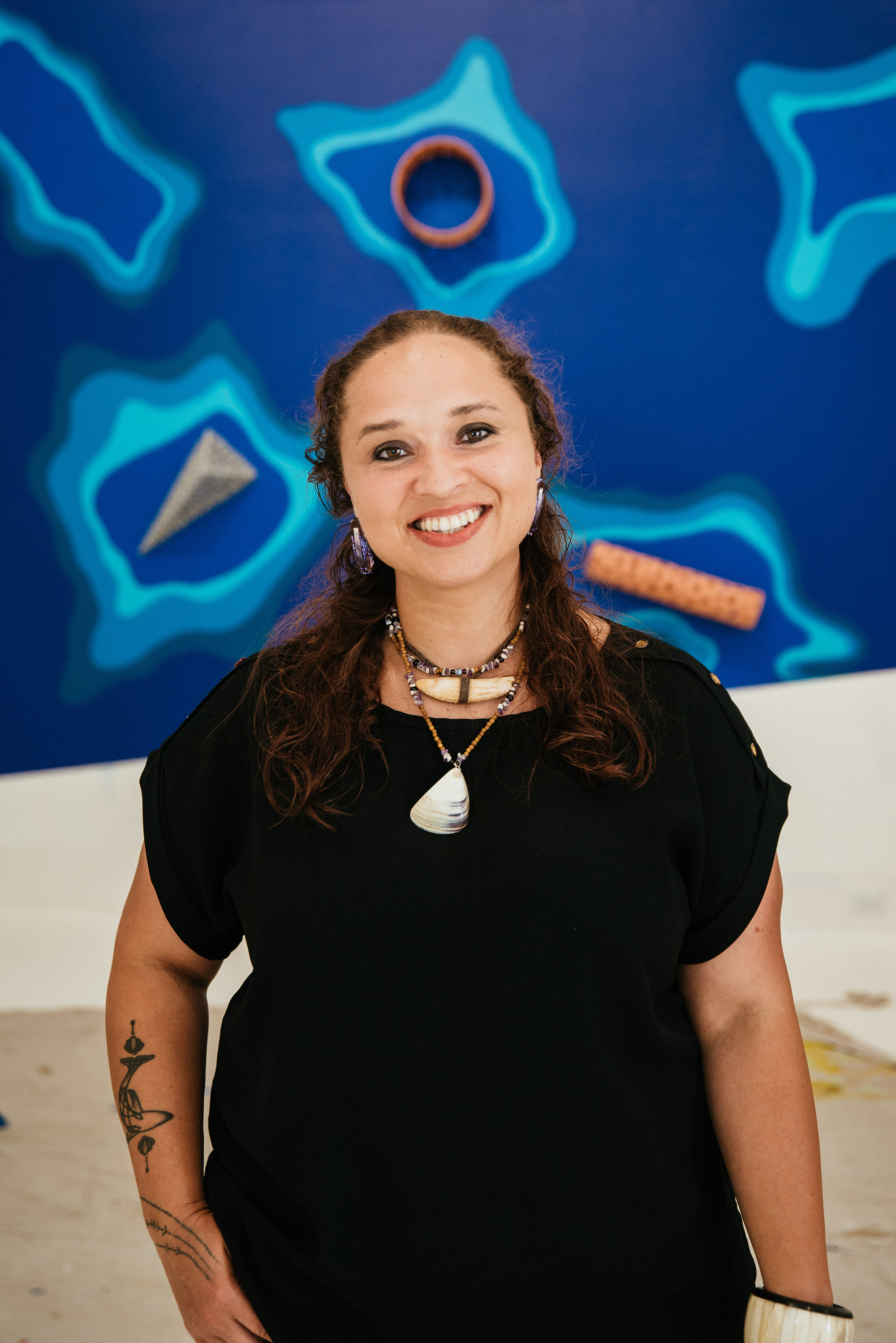 Indigenous woman standing in front of a blue background with objects attached to the wall. Woman is wearing a black shirt and has two necklaces on made of bone/shells. her hair is half up and she is smiling
