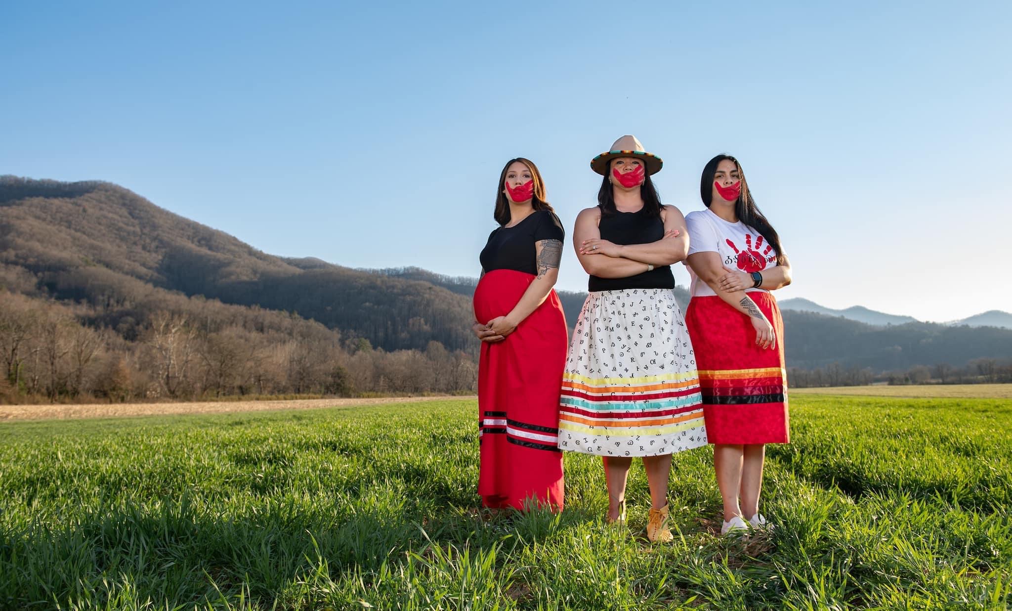 Three indigenous women standing near a mound dressed in red skirts with red handprints across the face