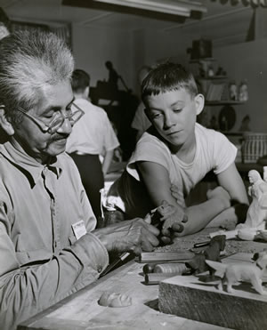A young boy looks on as Watty Chiltoskie demonstrates woodcarving. Southern Highland Craft Guild.
